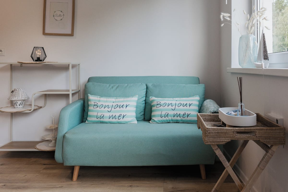 Ein blaugrünes Sofa mit zwei Kissen, ein kleines weißes Regal mit Dekoration und ein Korbtisch mit einer Schale und einem Reed-Diffuser.