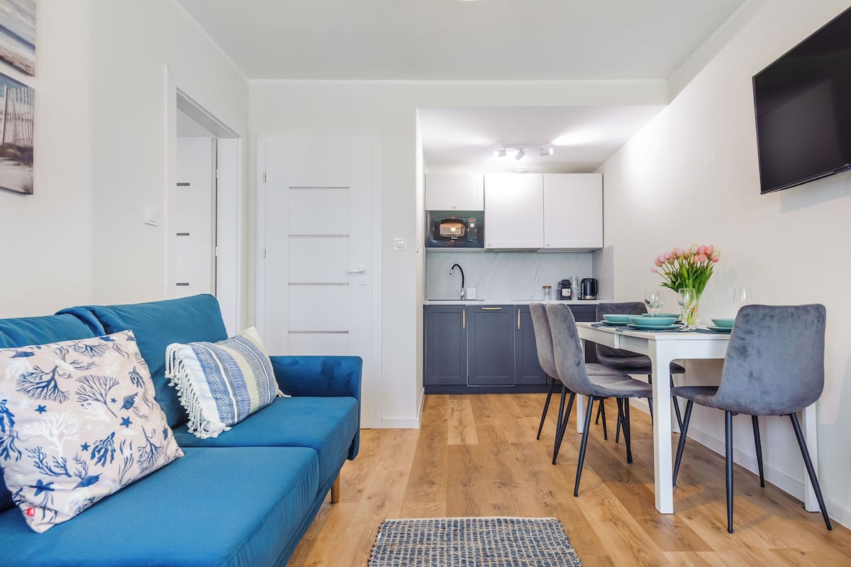 Ein kompakter Wohnbereich mit blauem Sofa, Esstisch mit Stühlen, Küchenzeile und einem an der Wand befestigten Flachbildfernseher.