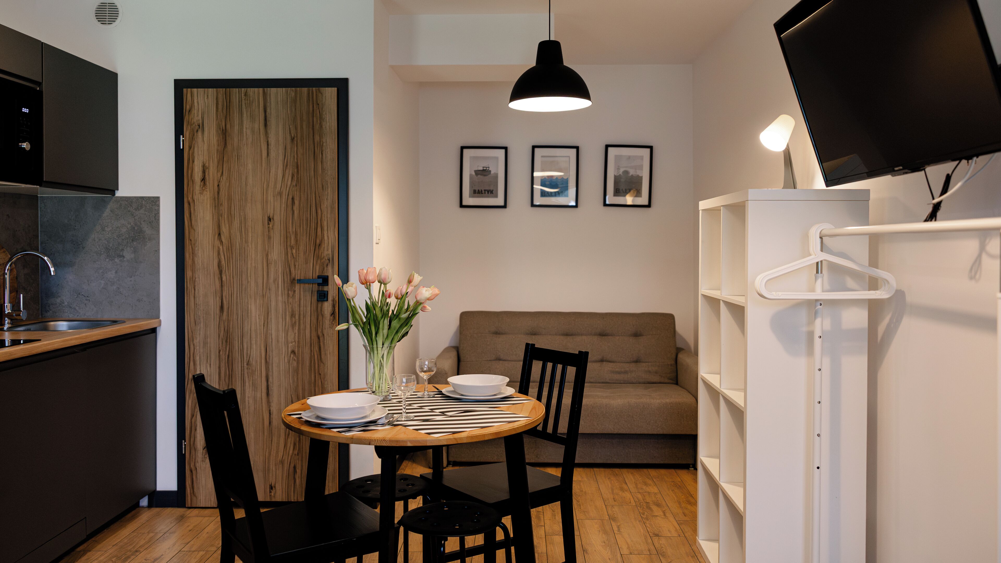 Una cocina compacta con puerta de madera, una mesa de comedor con sillas, un sofá blanco y un televisor montado en la pared.