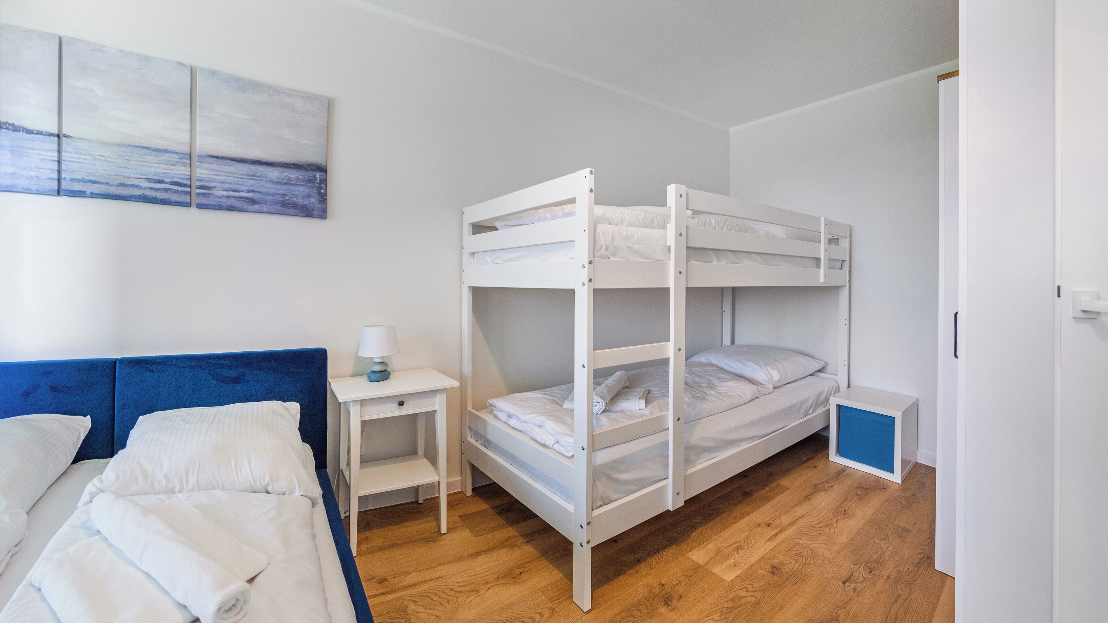 Habitació amb lliteres, un panell blau al capçal, una taula de nit amb una làmpada i un quadre a la paret.