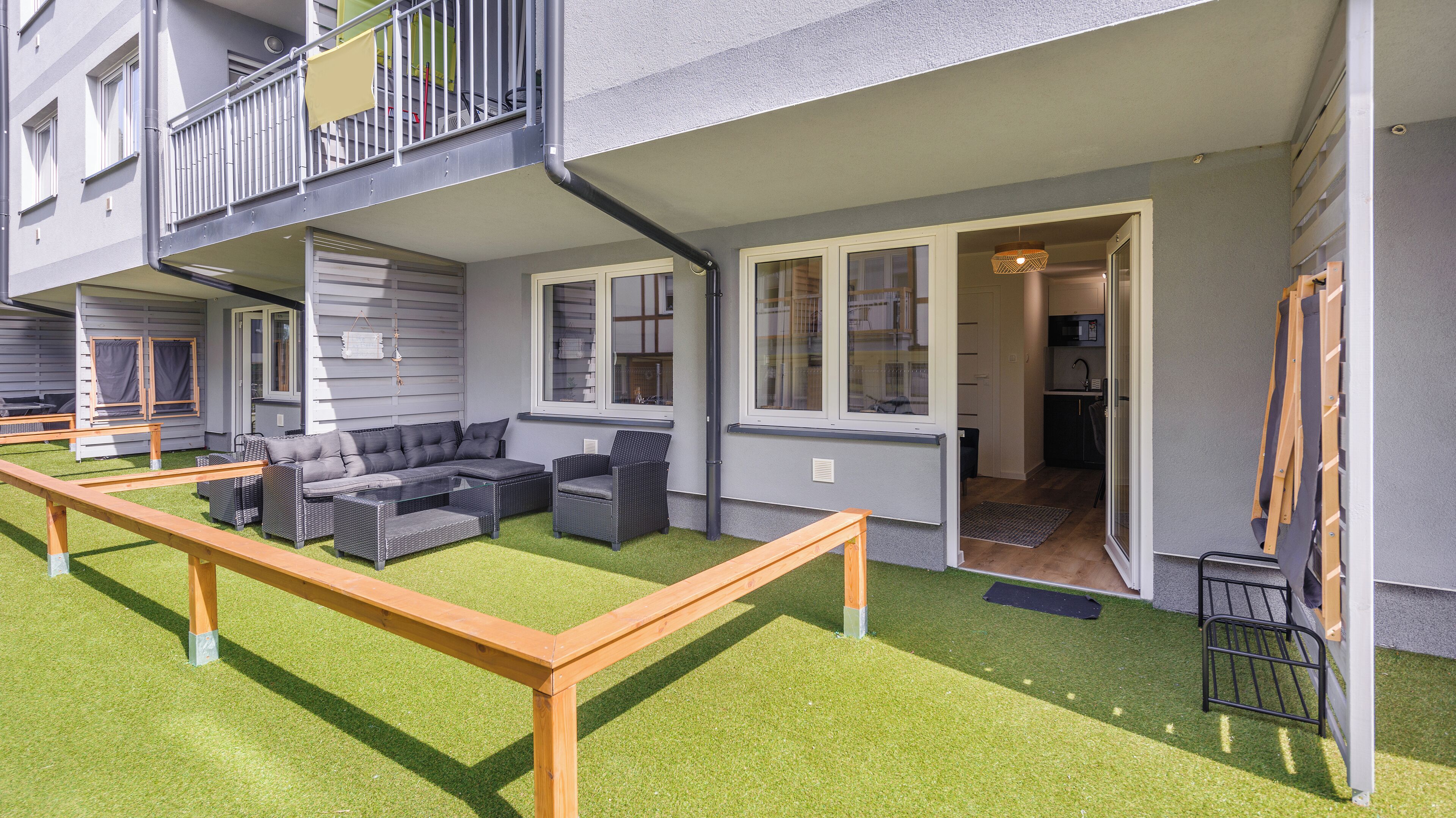 A modern apartment with a balcony, a living area with a sofa and chairs, and a small kitchen visible through an open door.