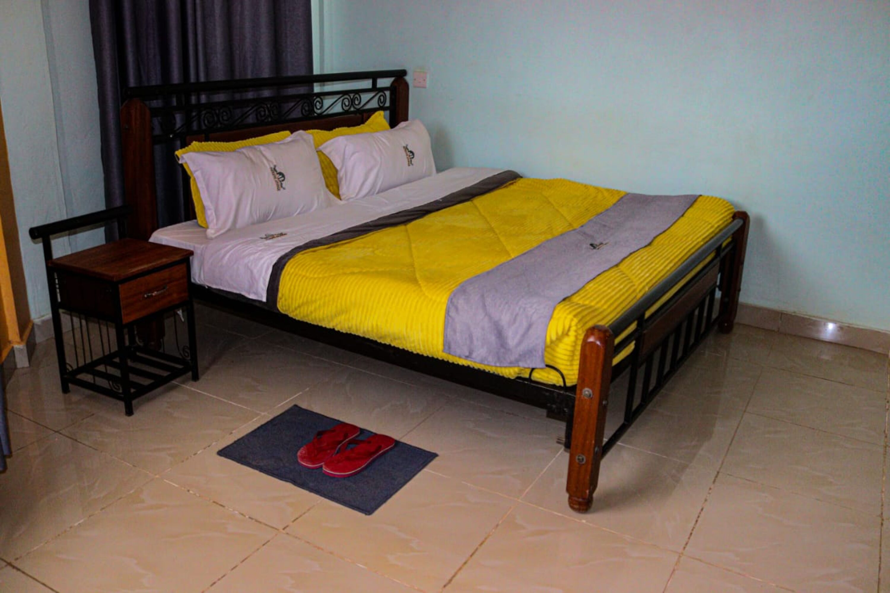 A bed with a wooden frame, a yellow and purple bedspread, and a pair of red slippers on a blue mat.
