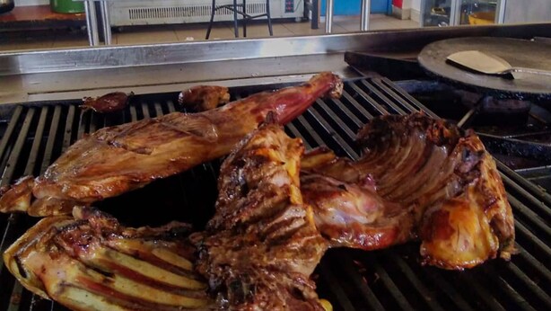 Una parrilla con costillas asándose. Al fondo, se observa un mostrador con una caja registradora, una silla y un bote de basura.