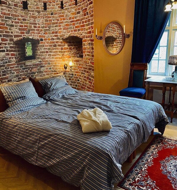 Un dormitorio con pared de ladrillo, una cama con ropa de cama a rayas, una mesita con una lámpara y una ventana con cortinas.
