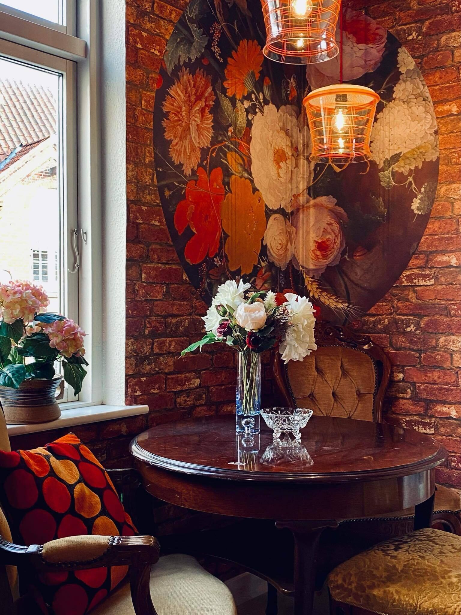 Una habitación con pared de ladrillo, una mesa redonda de madera con un florero, una silla con cojín estampado y un tapiz floral grande.