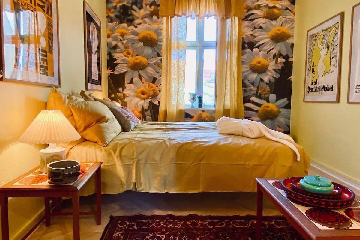 Une chambre à coucher comprenant un lit, une table de chevet, une lampe, une fenêtre avec des rideaux et une décoration murale à motifs floraux.
