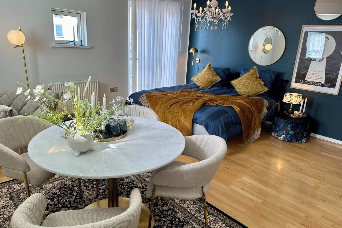 Un salon moderne avec une table ronde, deux fauteuils, un canapé et un lit doté d’une tête de lit bleue.