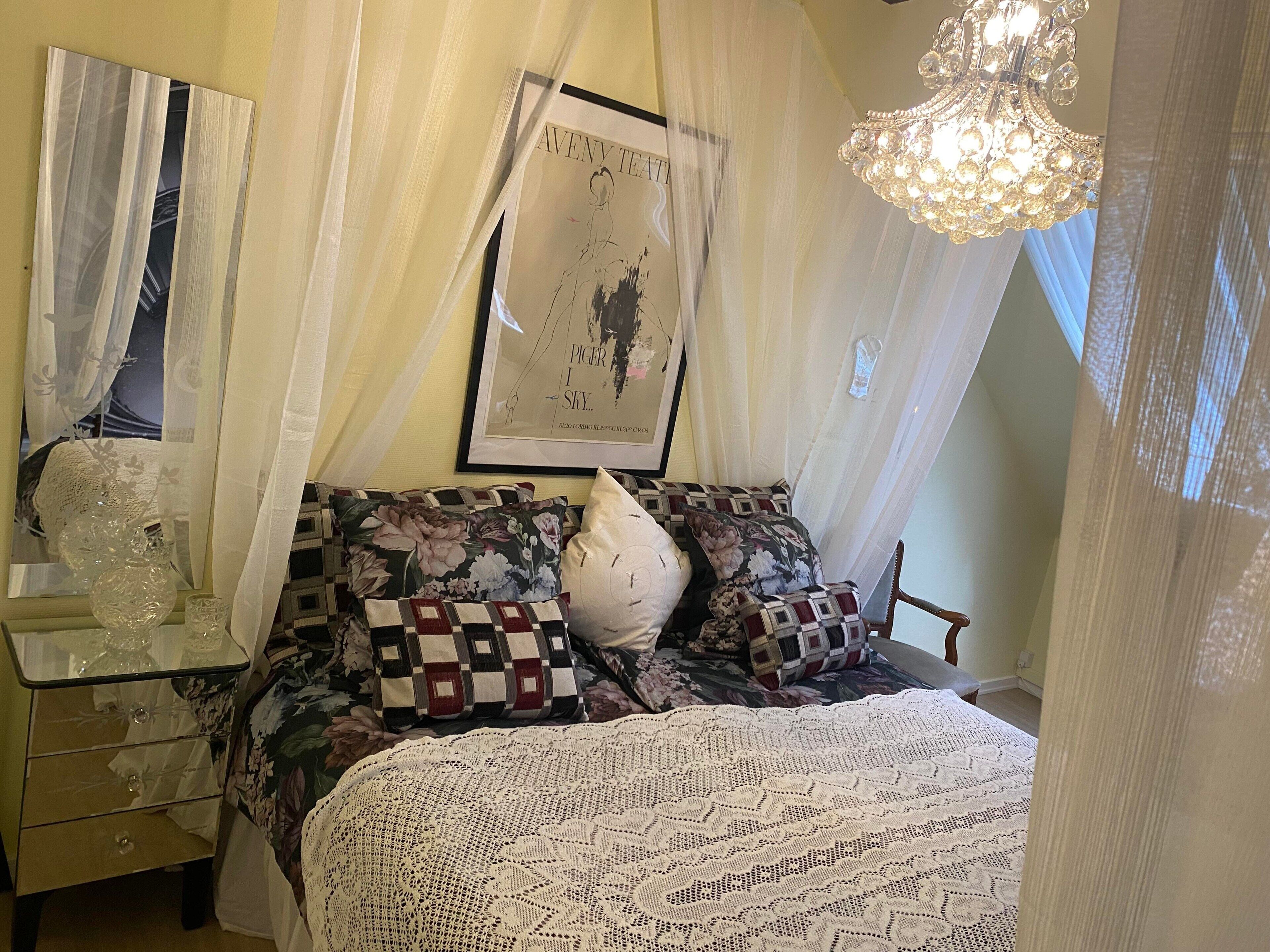 A bedroom with a bed, a nightstand, a mirror, a chandelier, and a framed poster on the wall.