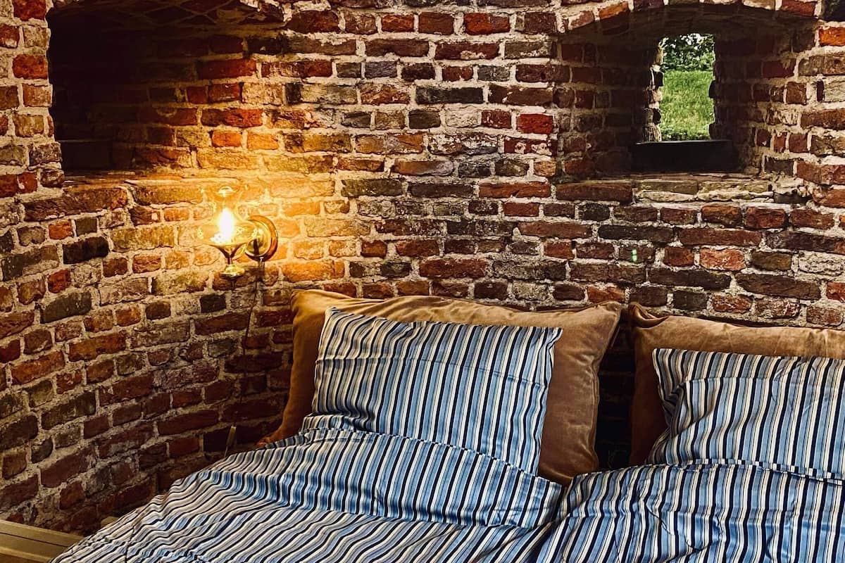 Une pièce avec un mur en briques, une applique murale allumée et un lit recouvert d’une literie à rayures.