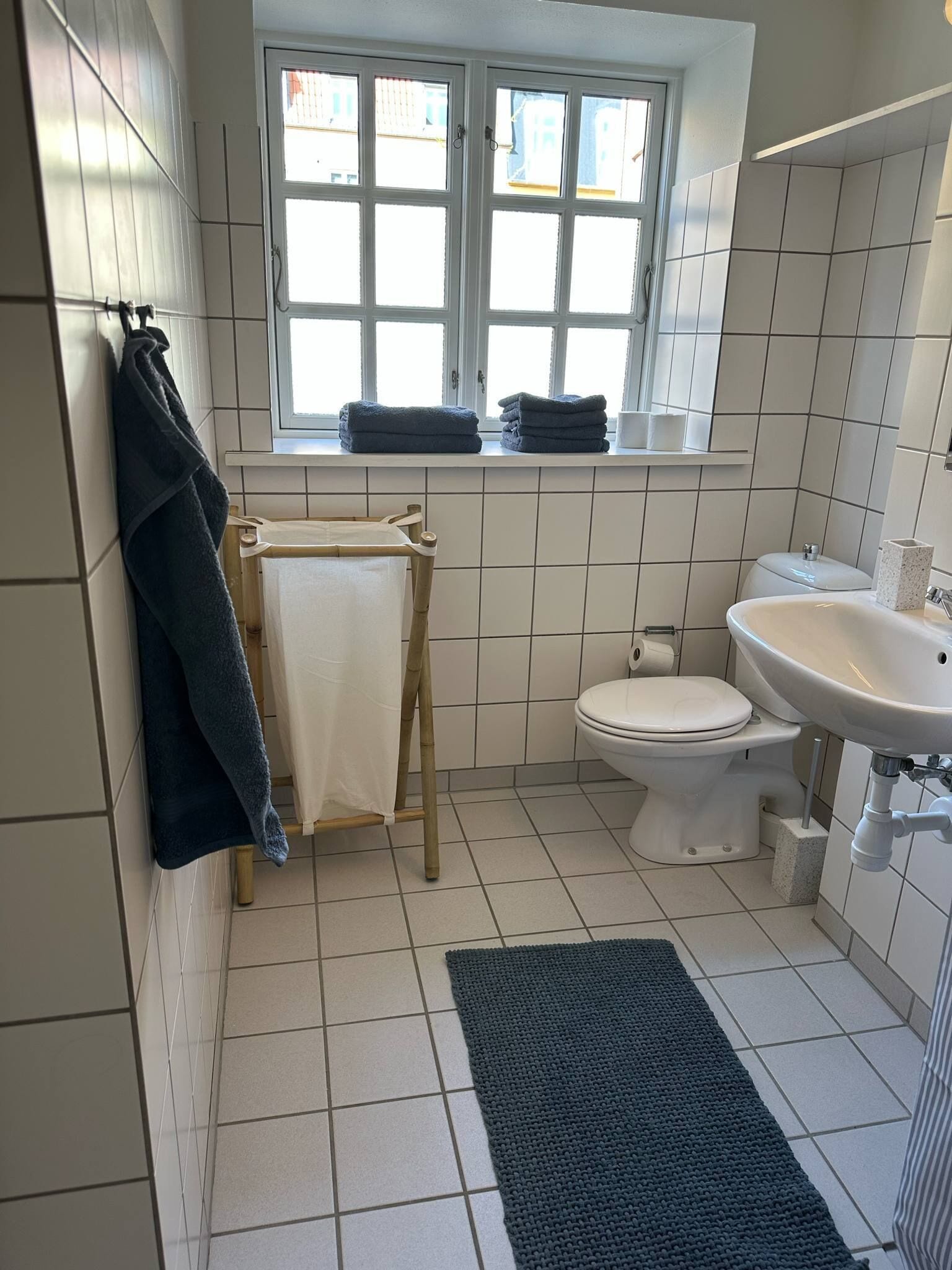 Une petite salle de bain propre, avec des carreaux blancs, des toilettes, un lavabo, une fenêtre et un porte-serviettes.