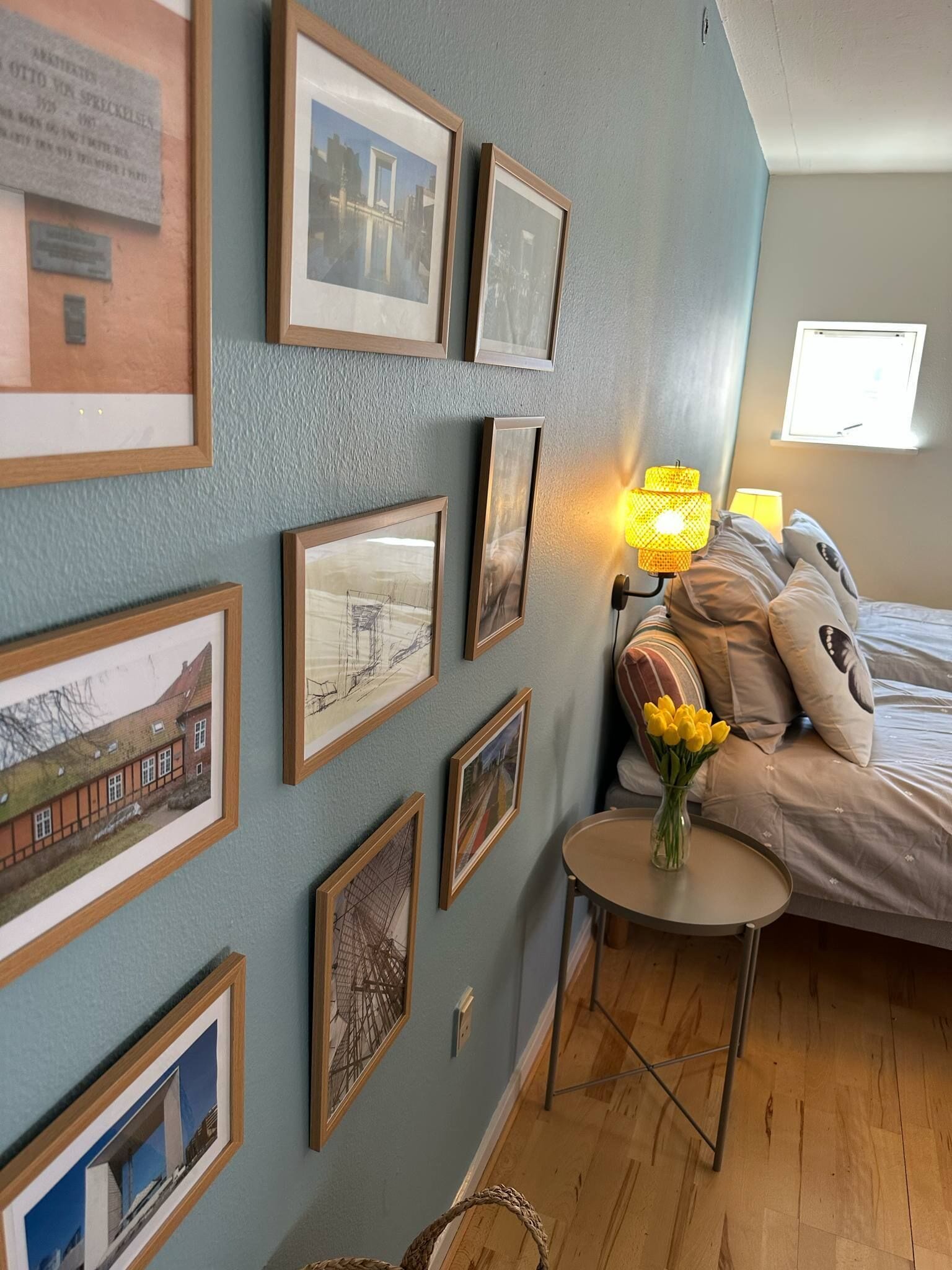 Une chambre à coucher avec un lit, une table de chevet avec une lampe, une petite table ronde ornée d’un vase de fleurs et plusieurs tableaux encadrés accrochés au mur.