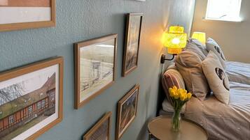 Une chambre à coucher avec un lit, une table de chevet avec une lampe, une petite table ronde ornée d’un vase de fleurs et plusieurs tableaux encadrés accrochés au mur.