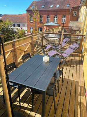 Una terraza de madera con una mesa, sillas, barandilla de vidrio y un edificio al fondo.