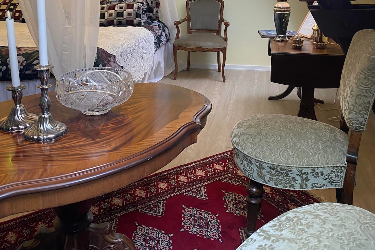 Une chambre à coucher avec un lit à baldaquin, une table en bois surmontée d’un bol en verre, une chaise, une lampe et un tapis rouge à motifs.