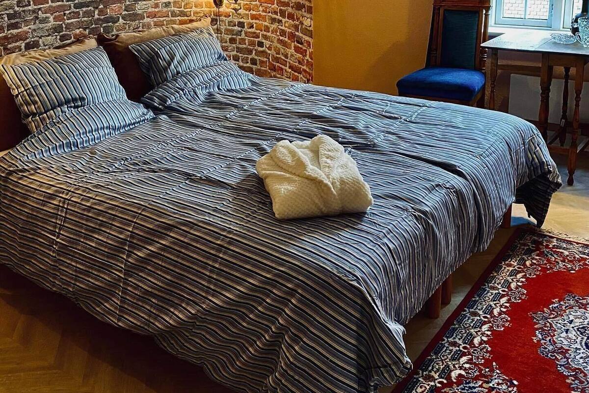 Une pièce avec un mur en briques, un lit avec une literie à rayures, une petite table avec une lampe et une fenêtre avec des rideaux.