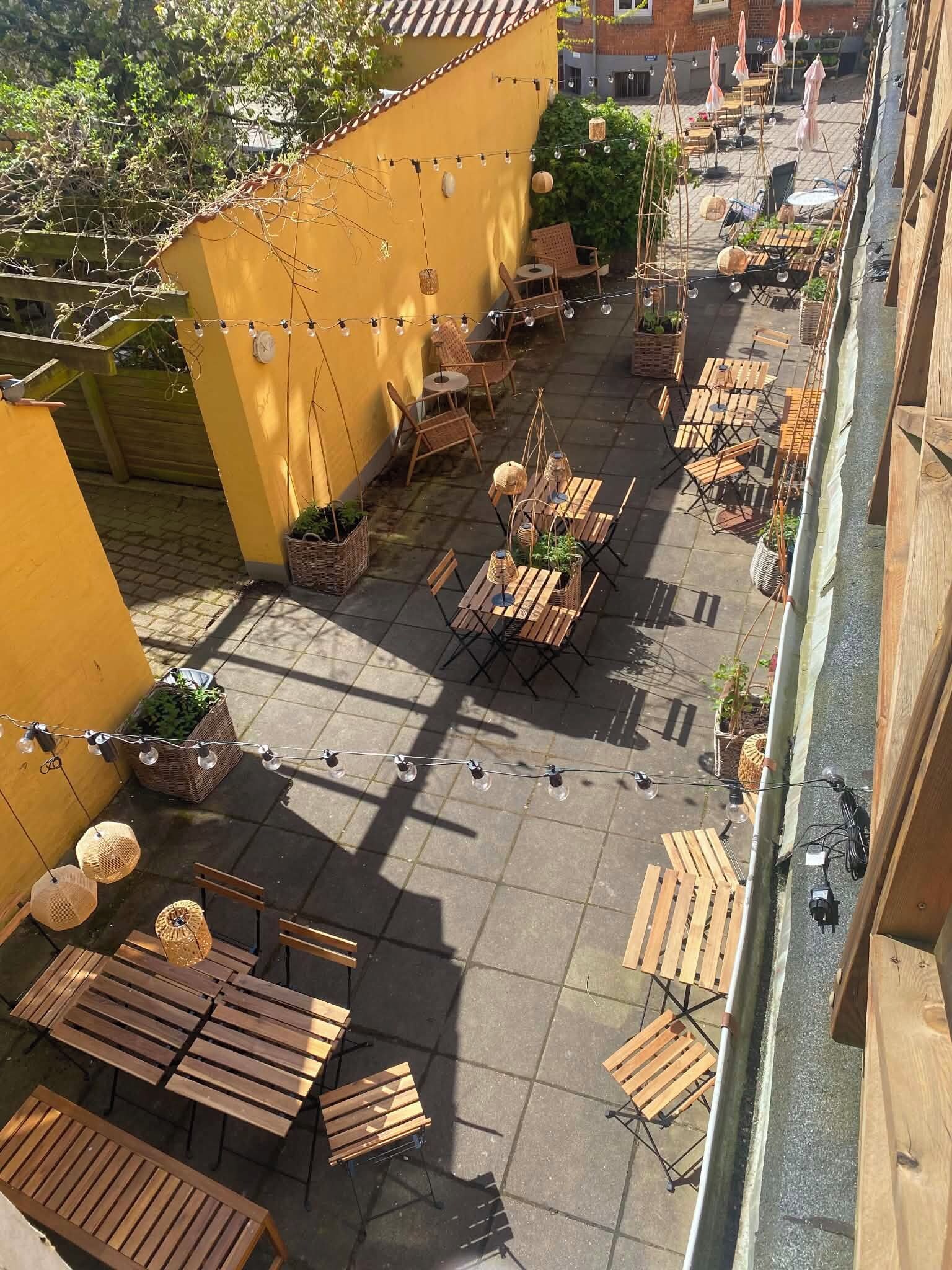An outdoor patio area with multiple wooden tables and chairs, string lights, and potted plants.