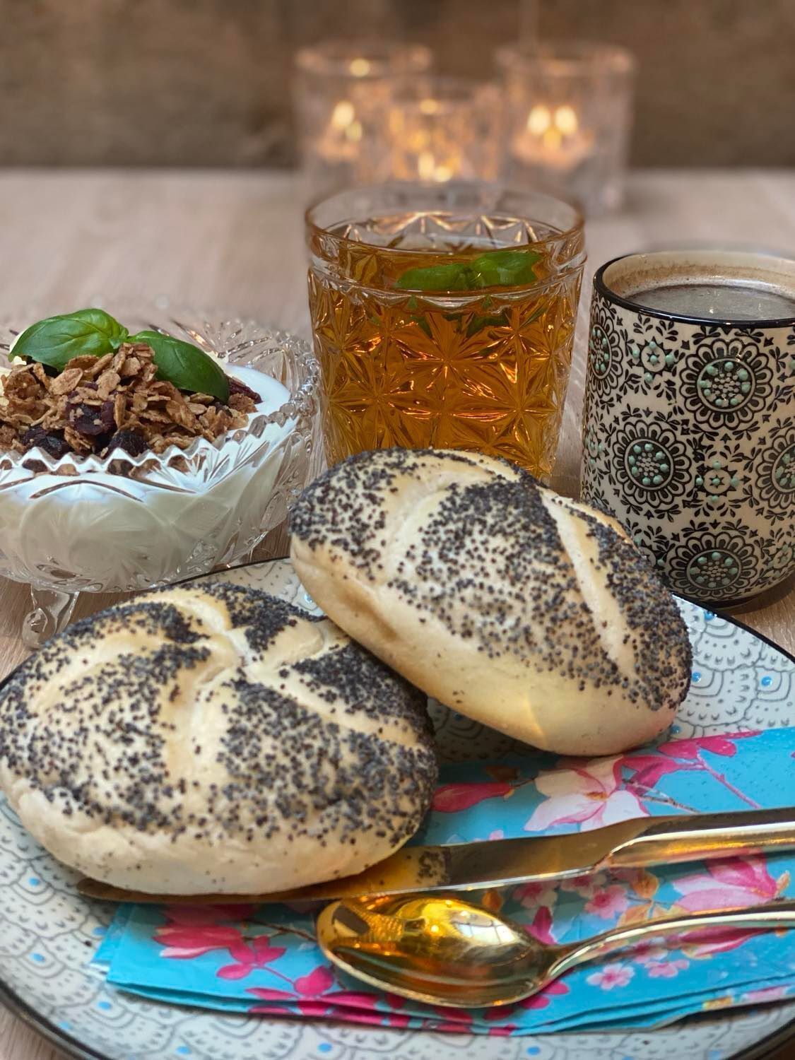 A plate with poppy-seeded bread, a bowl of granola with a basil leaf, a glass of tea, and a patterned mug.