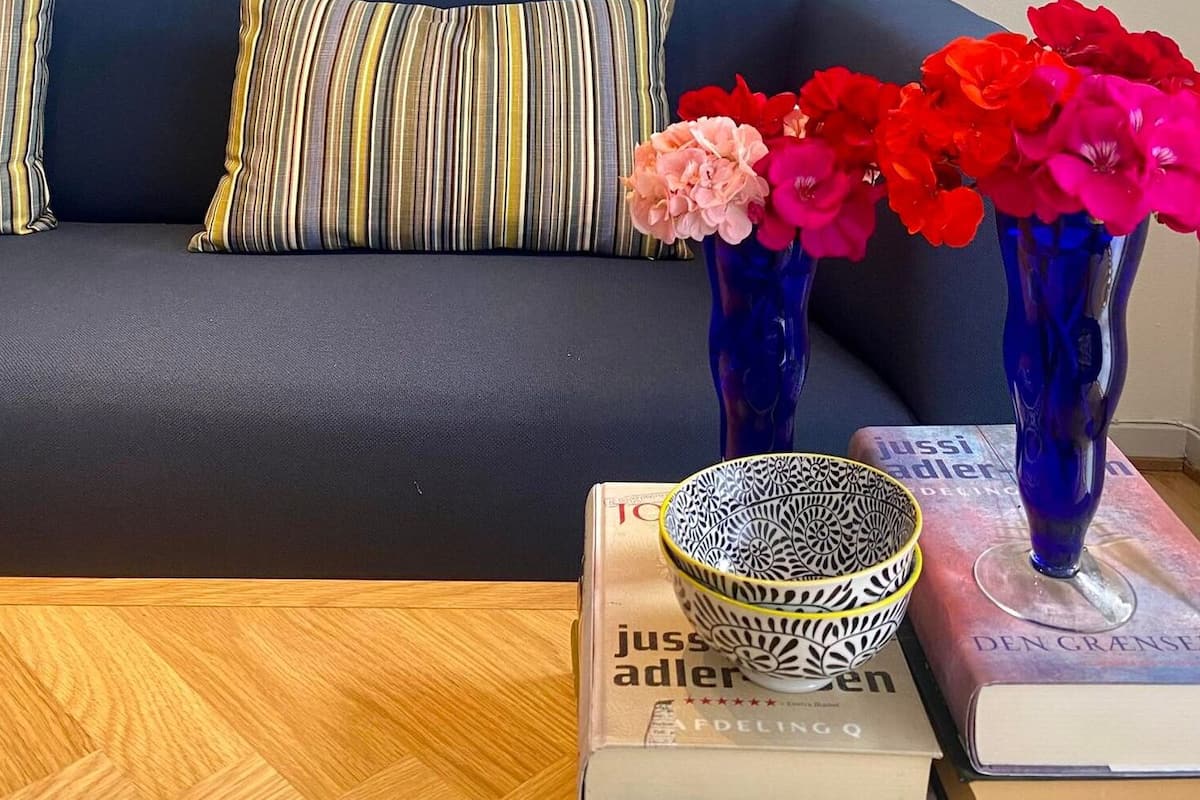 Un canapé agrémenté de coussins à rayures, un vase bleu rempli de fleurs et un bol à motifs posé sur une table où se trouvent des livres.