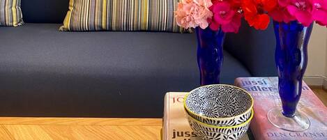 A sofa with striped pillows, a blue vase with flowers, and a patterned bowl on a table with books.