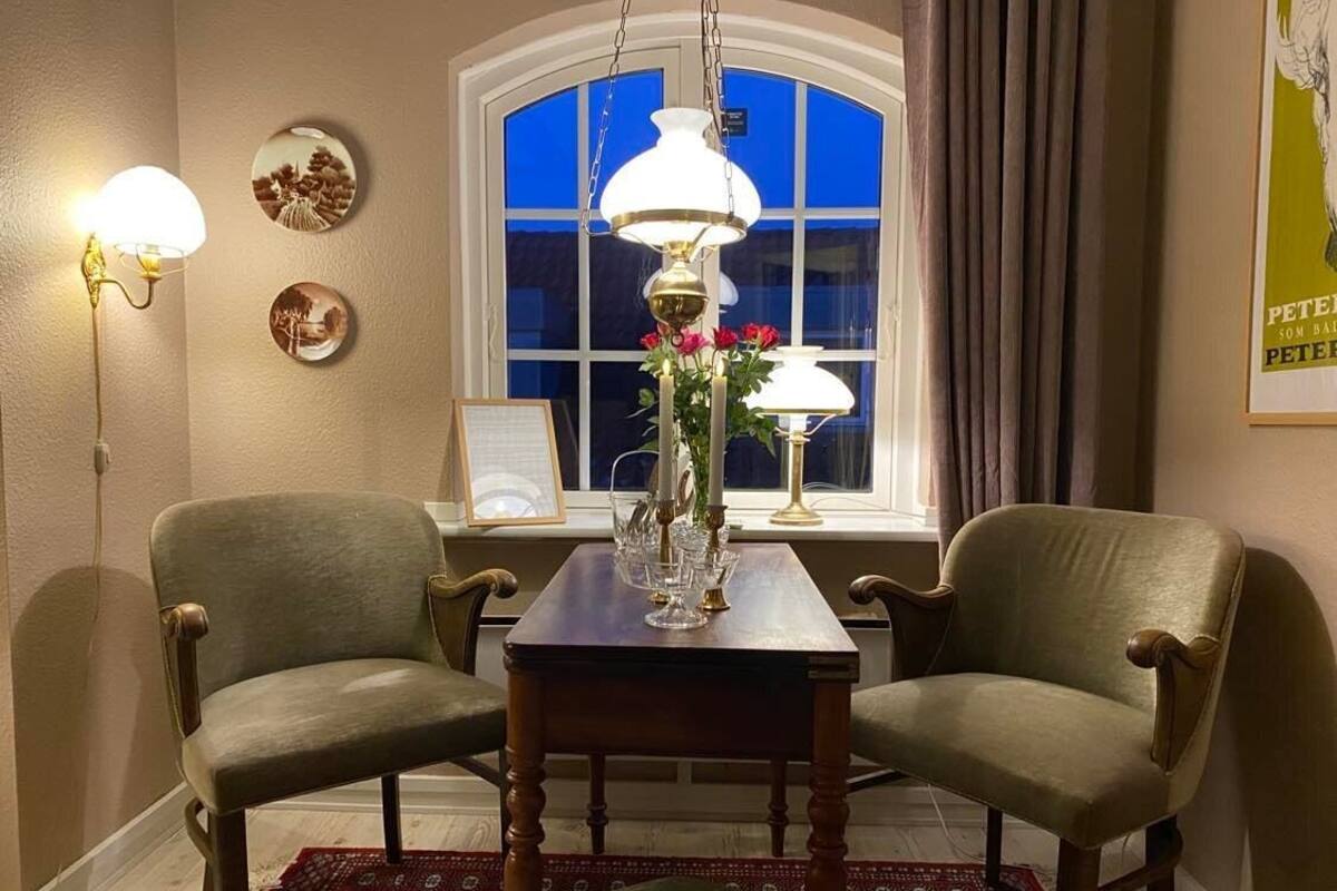 Un coin repas comprenant deux chaises, une table et un vase de fleurs. Il y a une fenêtre avec des rideaux et une lampe fixée au mur.