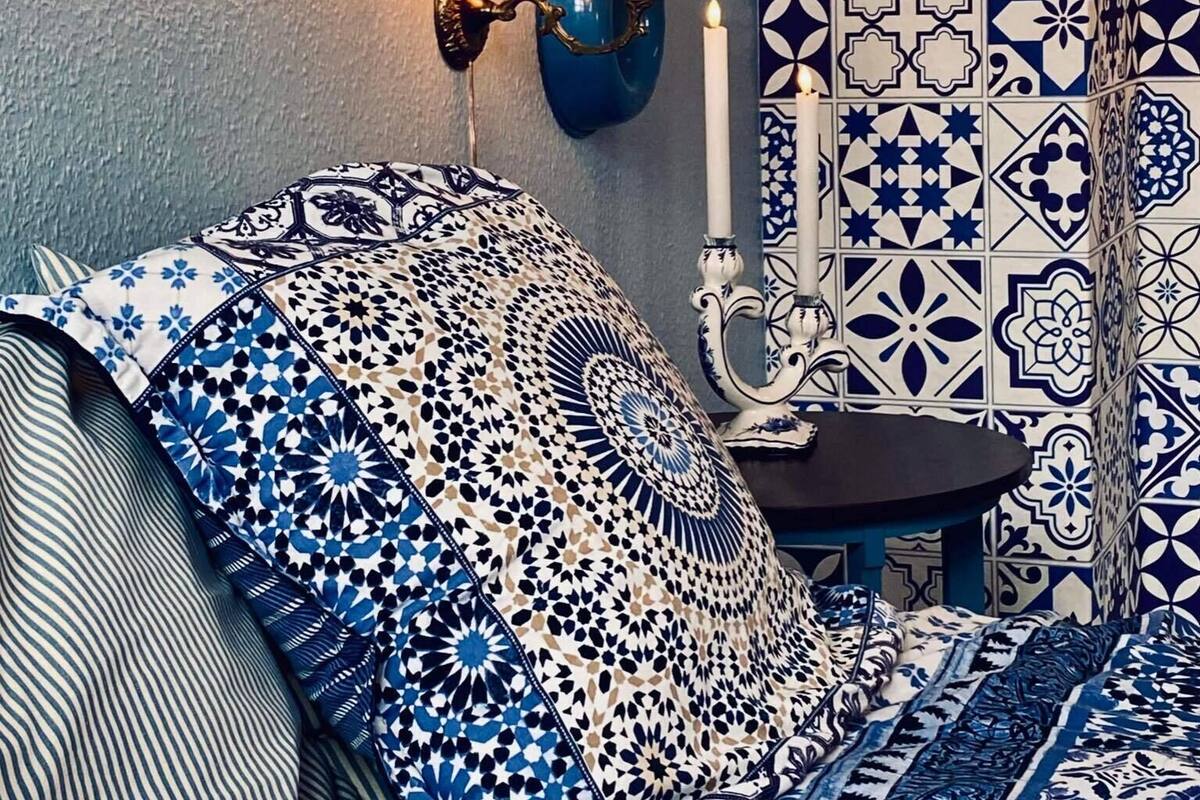 Une chambre avec une couvre-lit à motifs, une lampe fixée au mur et un mur recouvert de carreaux formant un motif géométrique.