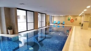 An indoor swimming pool with clear glass walls, blue mosaic tiles, and a few lounge chairs. - Damac Tower studio in the center of Amman (عمّان)