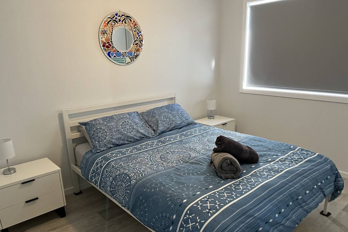 A neatly made bed with a blue patterned bedspread and pillows, a bedside table with a lamp, and a decorative mirror on the wall.