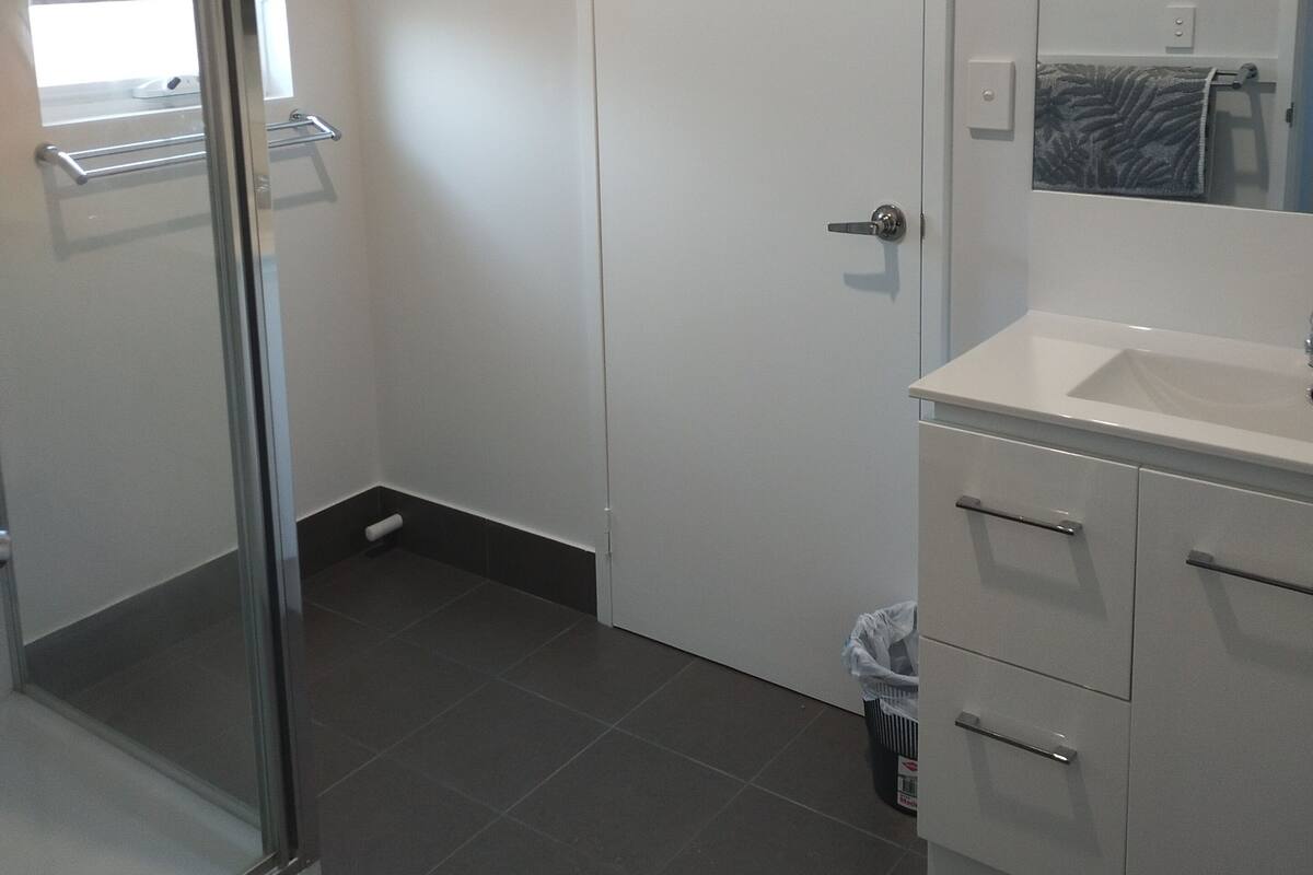 A bathroom with a glass shower door, a white door, a white sink with a mirror, and a trash bin.