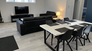 Una sala moderna con un sofá negro, un televisor, una mesa de comedor con sillas y una lámpara.