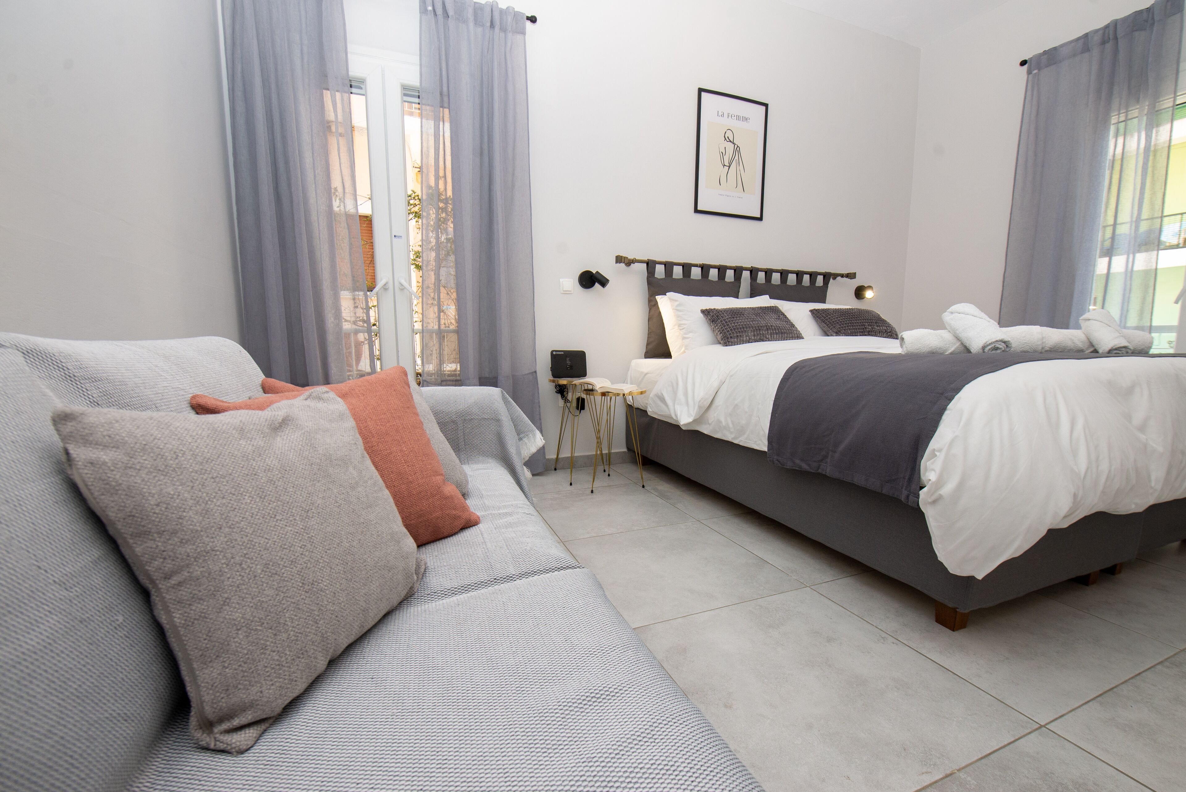 Ein modernes Schlafzimmer mit einem grauen Sofa, einem Bett mit weißer und grauer Bettwäsche und einem kleinen Tisch mit einer Lampe.