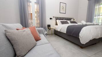 Ein modernes Schlafzimmer mit einem grauen Sofa, einem Bett mit weißer und grauer Bettwäsche und einem kleinen Tisch mit einer Lampe.