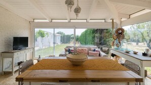 A spacious living area with a large wooden dining table, a TV on a white cabinet, and a view of the backyard through large windows. - Trullo Letizia con Piscina by Wonderful Italy (Ostuni)