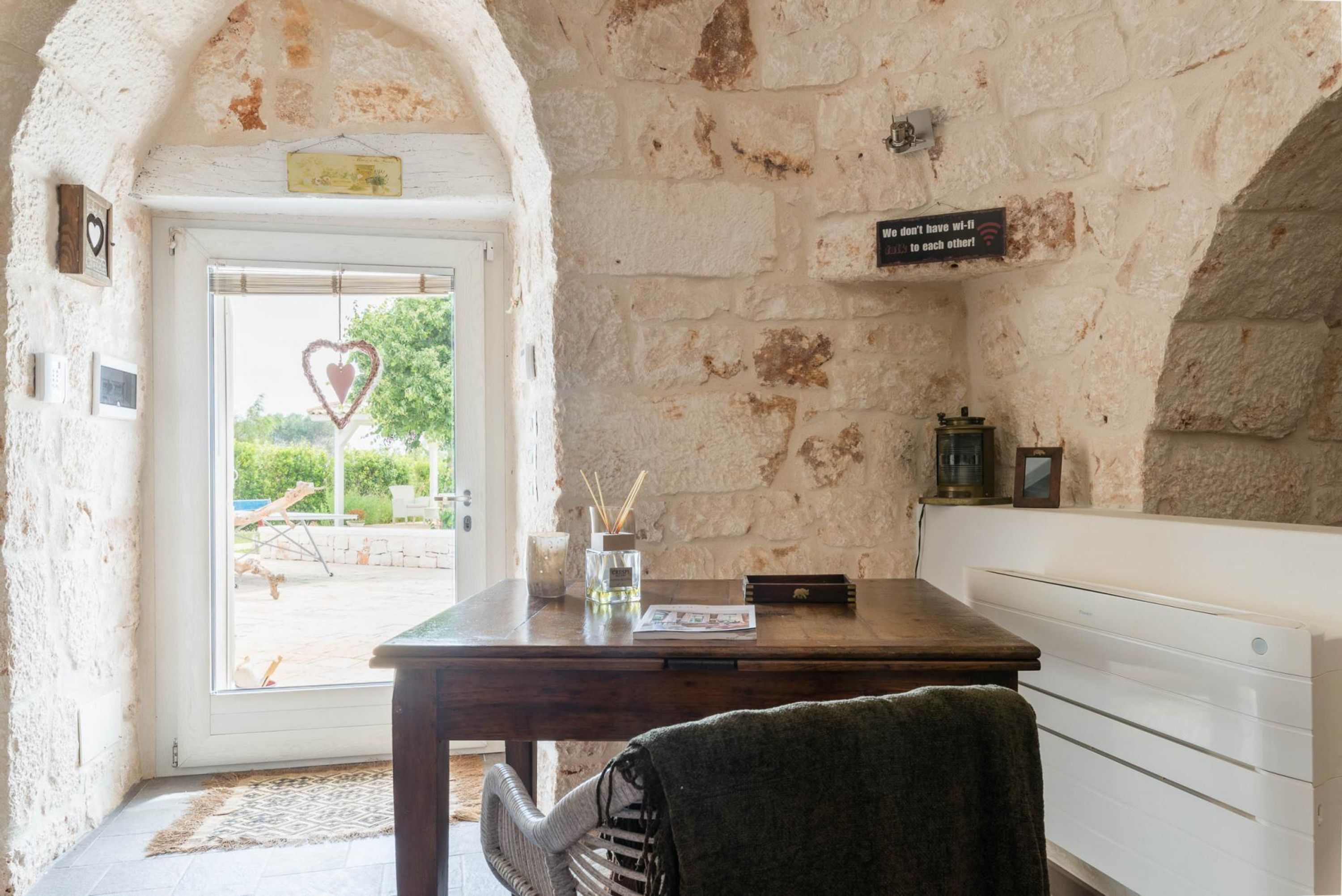 Una habitación rústica con paredes de piedra, un escritorio de madera, una silla y un banco. Una puerta da al exterior, y una decoración con forma de corazón cuelga en la pared.