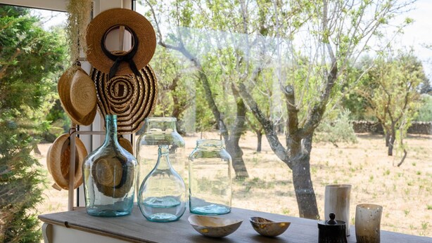 A table with woven hats, glass bottles, and ceramic items by a window overlooking a tree-lined area. - Trullo Letizia con Piscina by Wonderful Italy (Ostuni)