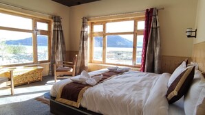 A bedroom with a large bed, a wooden desk, and a chair. There are large windows with curtains and a view of mountains. - Pangong Nest Cottage (Leh)