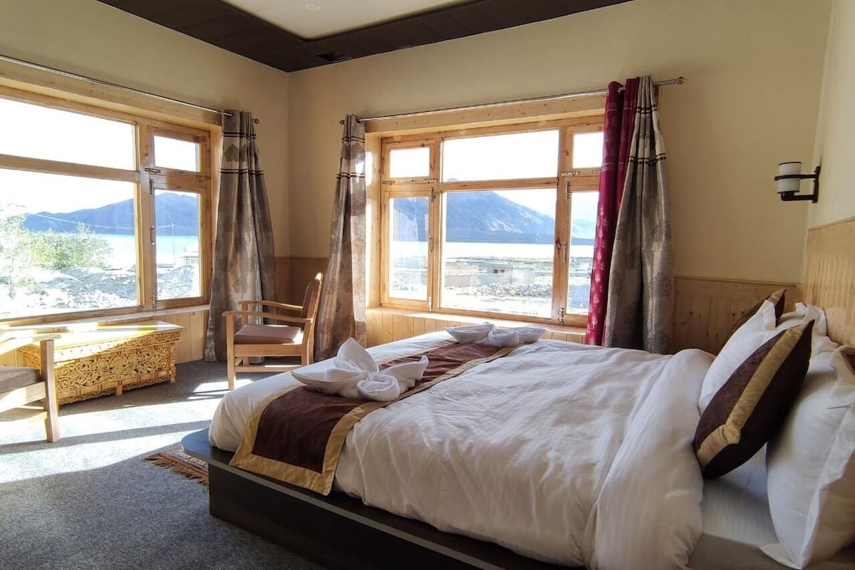 A bedroom with a large bed, a wooden desk, and a chair. There are large windows with curtains and a view of mountains.