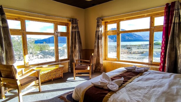 A room with a bed, a chair, and a table. There are large windows with curtains. - Pangong Nest Cottage (Leh)