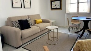 A living room with a beige sofa, a small round table, and a chair. There are two framed pictures on the wall. - 2 Flamenco AticoB (Cádiz)