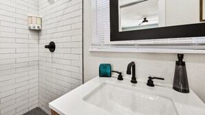 A bathroom with a white sink, black faucet, and a mirror above it. There is a shower area with white tiles and a handheld showerhead. - Quiet Studio Guesthouse in Jax - Near Downtown (Jacksonville)