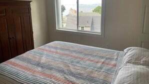 Une chambre avec une grande fenêtre, une armoire en bois et un lit recouvert d’une couette à rayures.