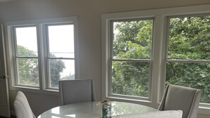 Un coin repas avec une table ronde en verre, entourée de quatre chaises grises à revêtement. Deux grandes fenêtres offrent une vue sur la verdure et un plan d’eau.