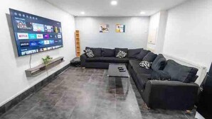 Un salon moderne doté d’un grand téléviseur à écran plat fixé au mur, qui diffuse différents services de streaming. On y trouve un canapé d’angle noir avec des coussins à motifs et une petite table basse au centre. Le sol est carrelé et les murs sont blancs.