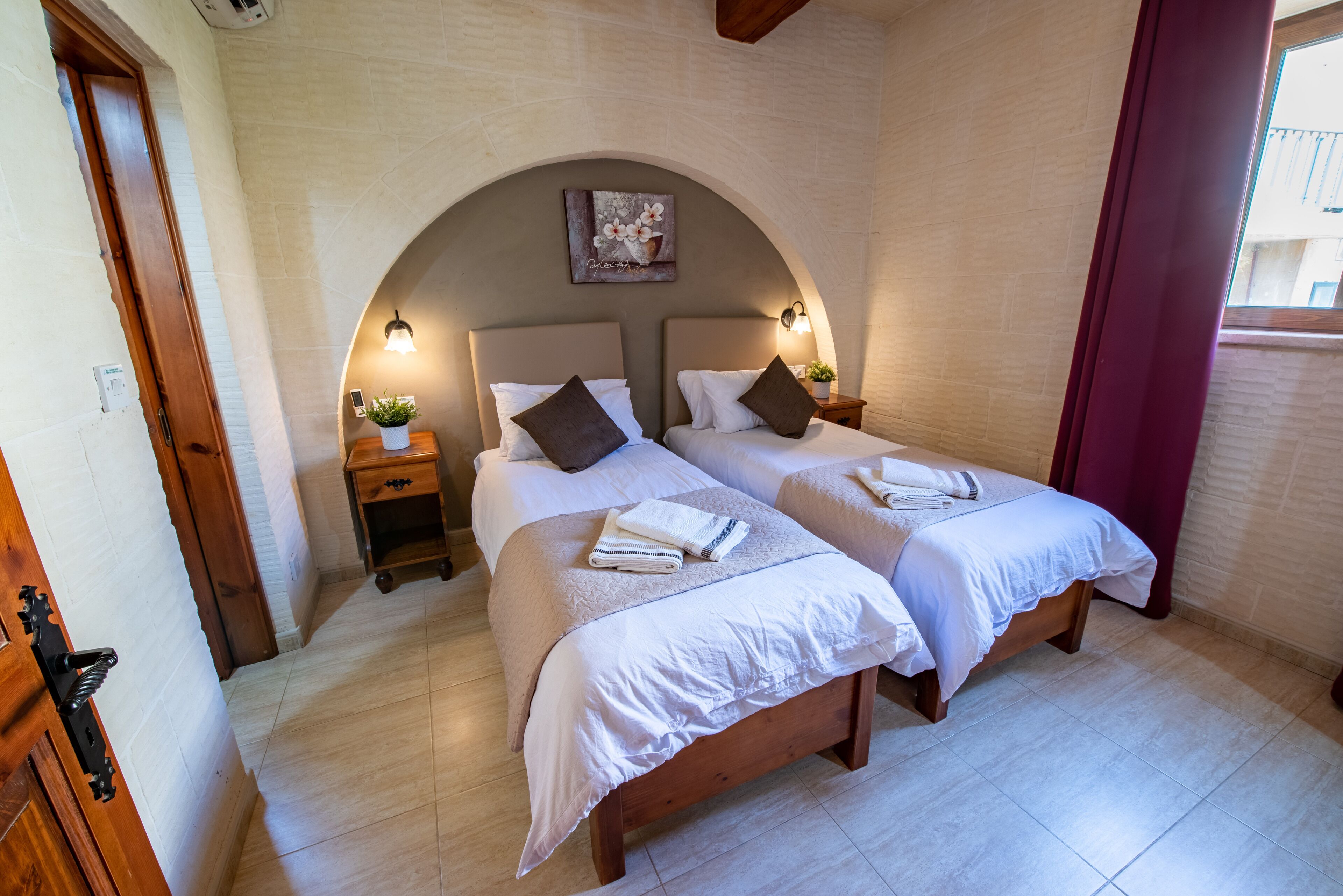 A double room with two beds, a small bedside table with a lamp, a wall-mounted artwork, and a window with curtains.