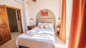 Un dormitorio con una cama grande, cabecera de madera, dos mesitas de noche con lámparas, aire acondicionado instalado en la pared y una puerta que da a otra habitación.