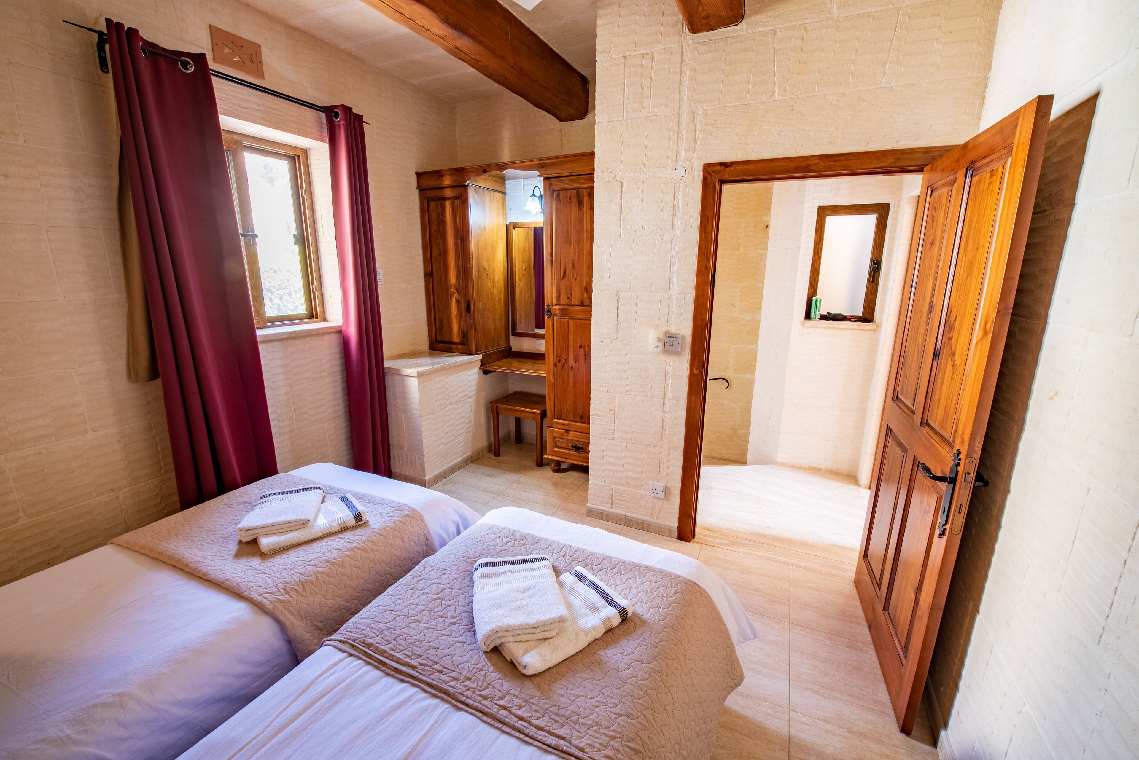 A hotel room with two beds, wooden furniture, a window with curtains, and a door leading to another room.