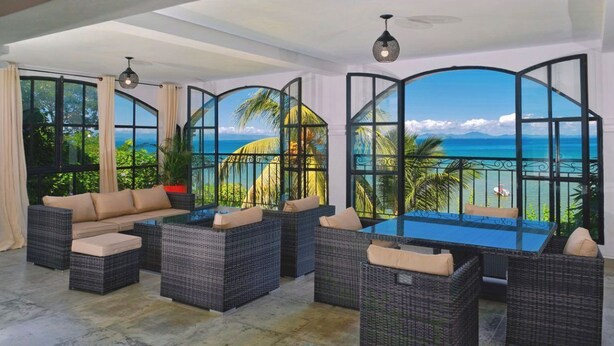 A spacious indoor area with a large window offering a view of the ocean and palm trees. There is a seating area with wicker furniture and a glass-top table. - Les Terrasses d'Ulysse (Nosy Be)