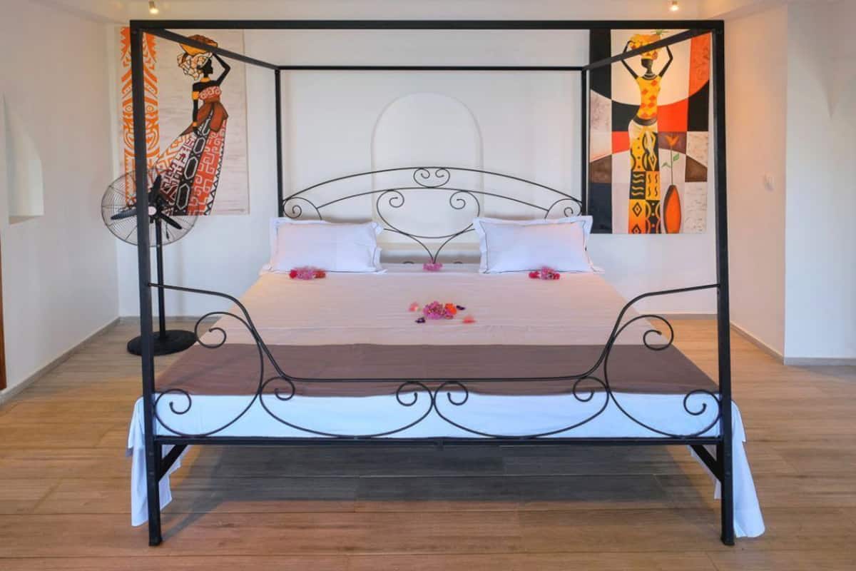 Una cama con dosel cubierta con una colcha blanca y flores rosadas, ubicada en una habitación con piso de madera y dos cuadros abstractos en las paredes.