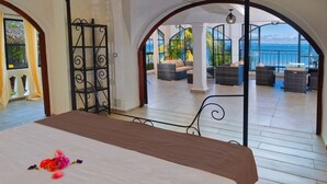 A hotel room with a large bed, a balcony with wicker furniture, and a view of the sea. - Les Terrasses d'Ulysse (Nosy Be)