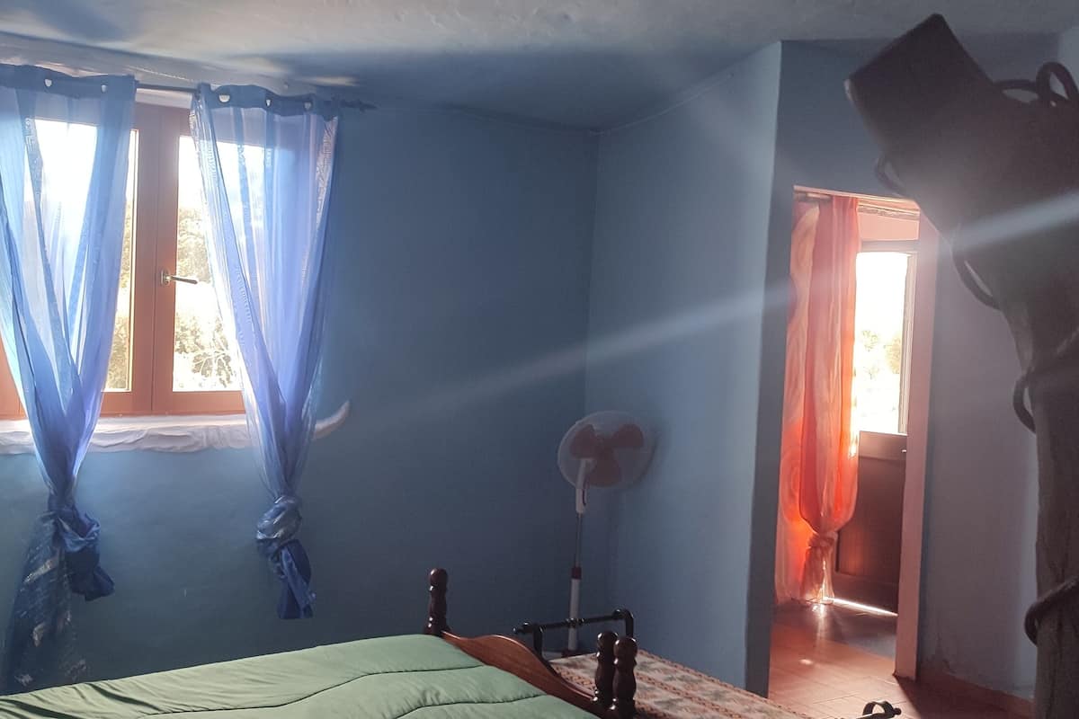 A room with a bed, a window with blue curtains, a fan, and a door leading to another room.