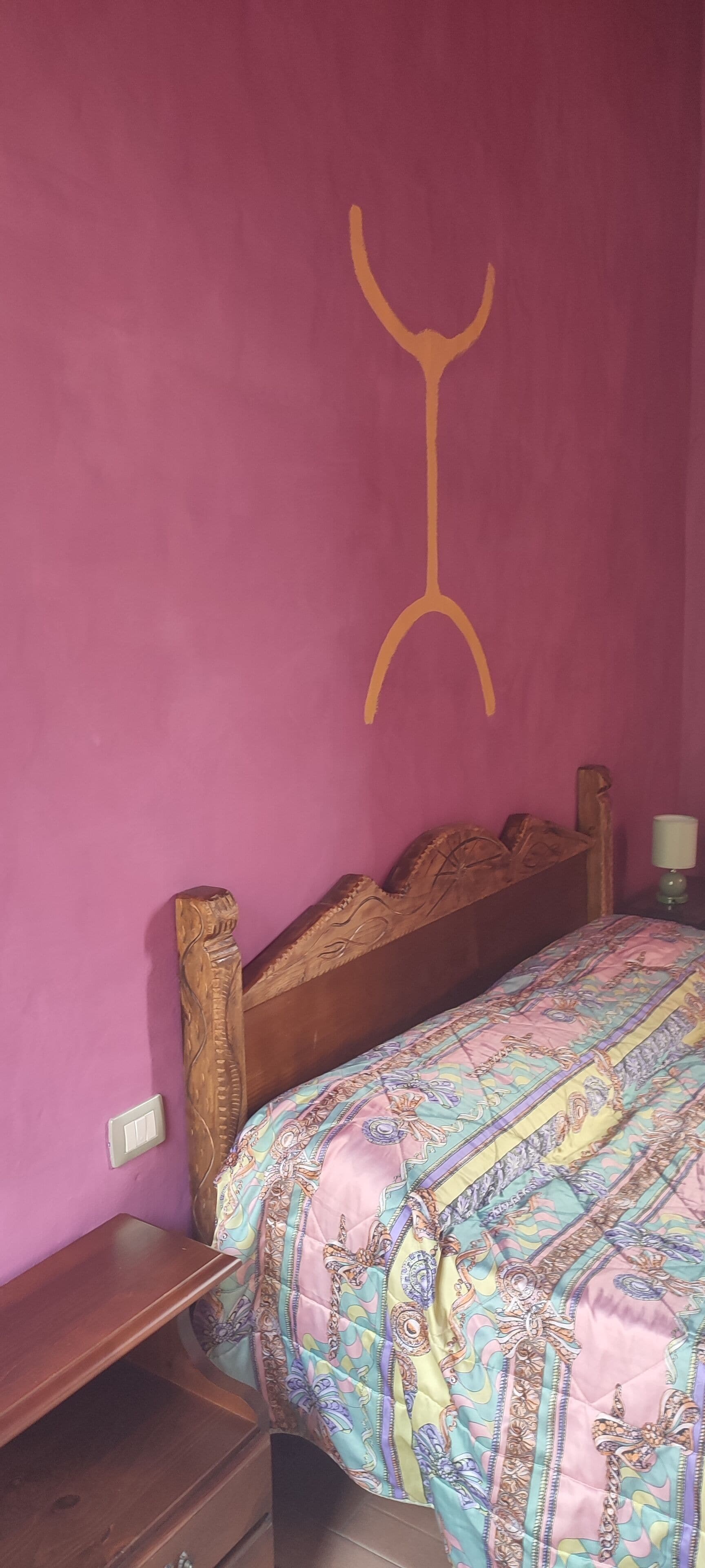 Una camera da letto con un letto in legno, una coperta a motivi decorativi, un comodino con una lampada e un punto luce a parete.